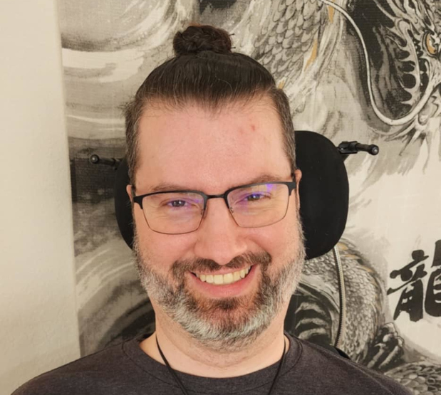 A white man's smiling face. He has brown hair pulled up in a bun a graybeard, and is wearing glasses.